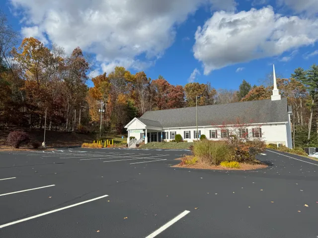 A church with a white exterior and tall steeple, surrounded by trees with fall foliage, and an empty parking lot in the foreground.