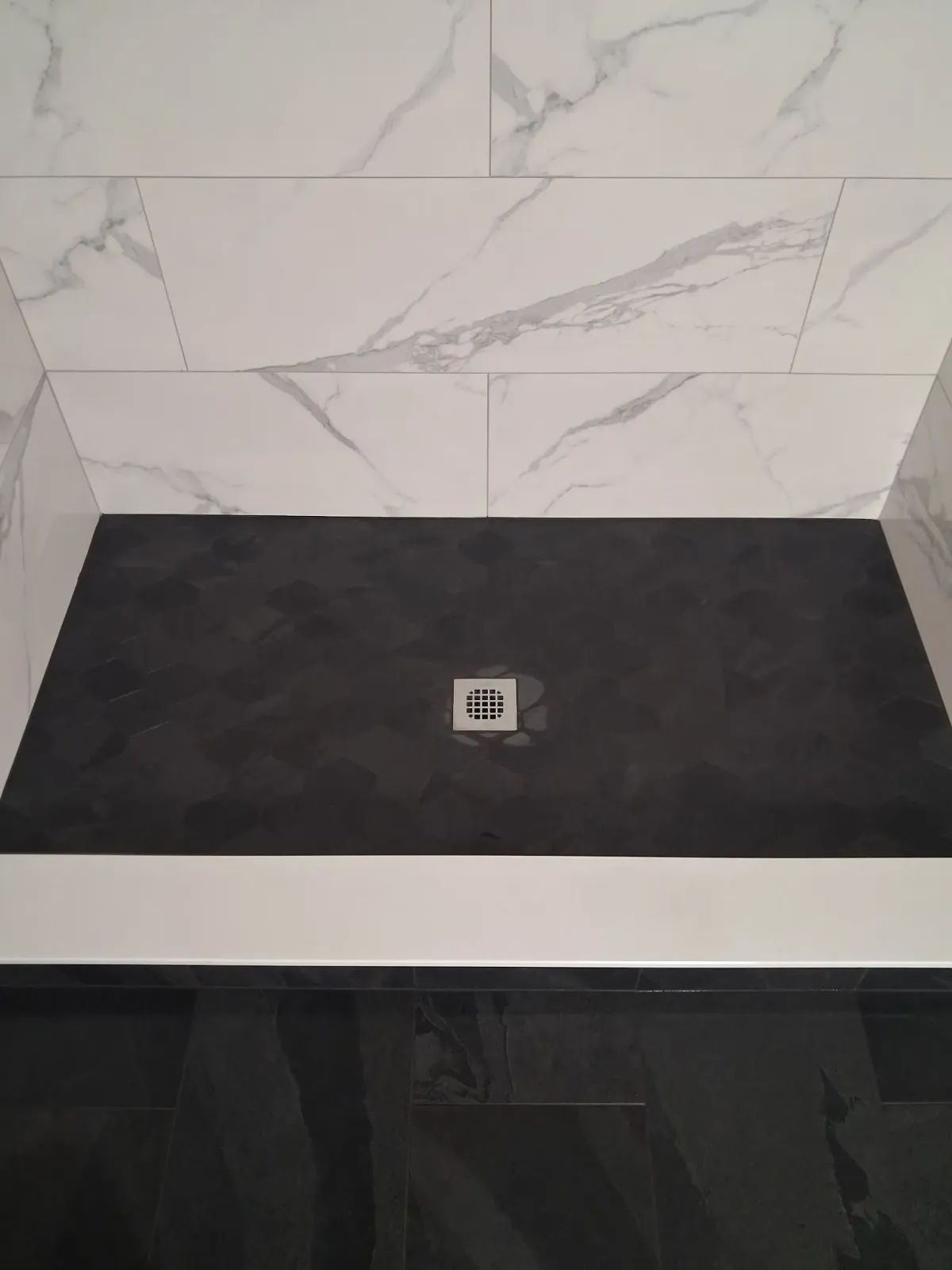 Empty shower base with black floor, white marble-pattern wall tile, and a centered drain.