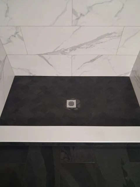 Empty shower base with black floor, white marble-pattern wall tile, and a centered drain.
