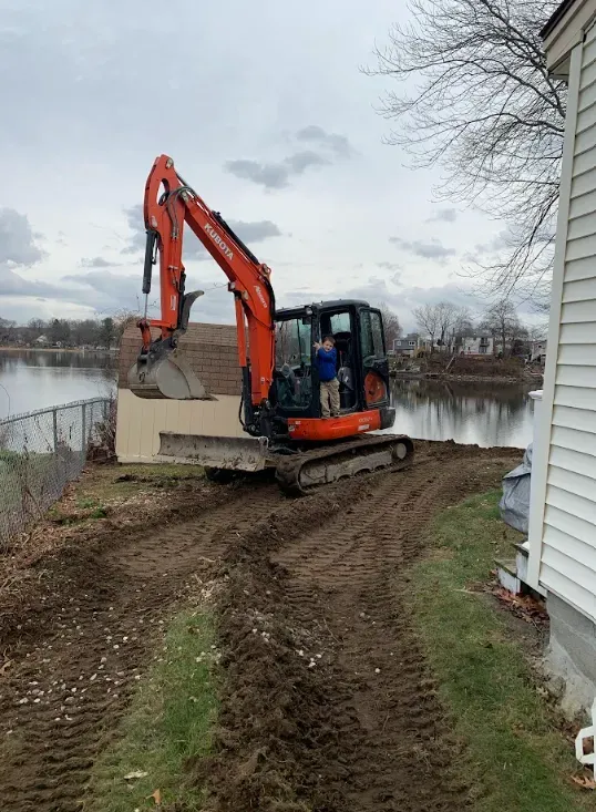Orange excavator working beside a house near water on a muddy path