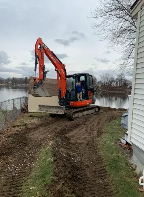 Orange excavator working beside a house near water on a muddy path