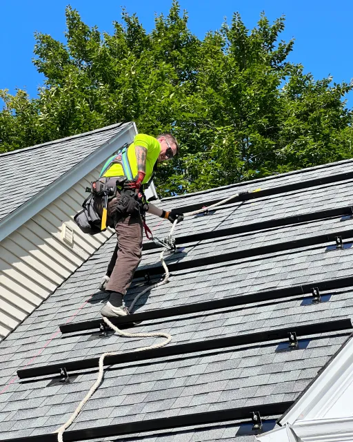 A worker in safety gear cleaning or inspecting a sloped roof with solar mountings during daytime, with a large tree and clear blue sky in the background.