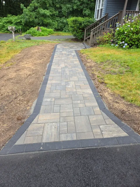 Stone walkway leading from a road to a house, bordered by mulch and green shrubs.