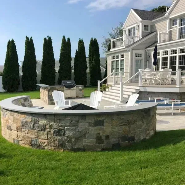 Stone fire pit with white chairs, outdoor kitchen, and a large house on a sunny day.