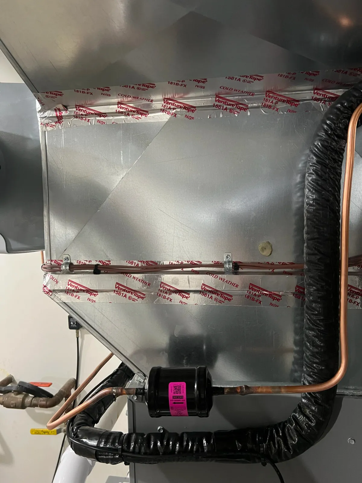 Close-up image of a metallic HVAC unit with various copper pipes, black insulated hose, and red and white VentureTape labels.