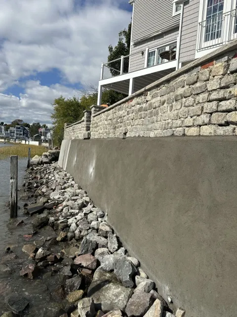 A waterfront retaining wall made of concrete and stone, with a house on top. Rocks line the water's edge.