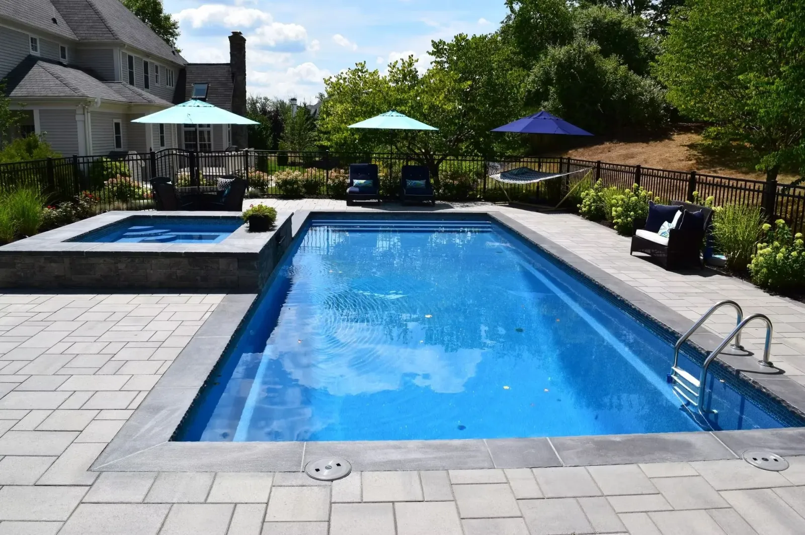 Swimming pool and hot tub with lounge chairs and umbrellas in a backyard. Blue water, sunny day.