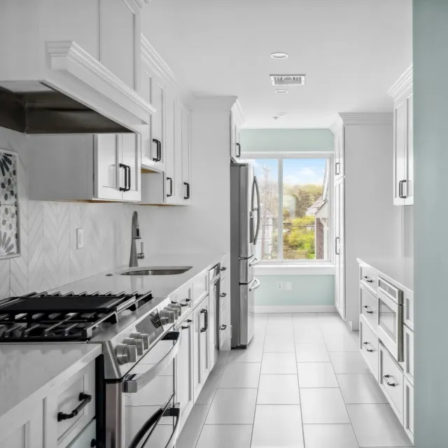 A modern kitchen with white cabinets, stainless steel appliances, tiled floor, and a large window overlooking a neighborhood with trees and houses.