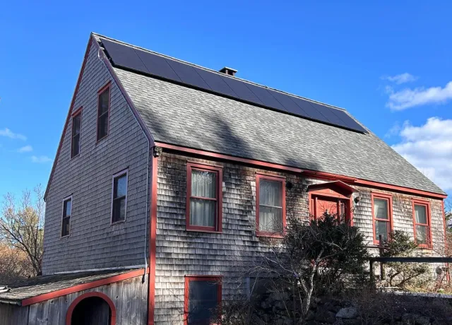 A two-story house with weathered wooden siding, red window trims, solar panels on the roof, and a clear blue sky in the background.