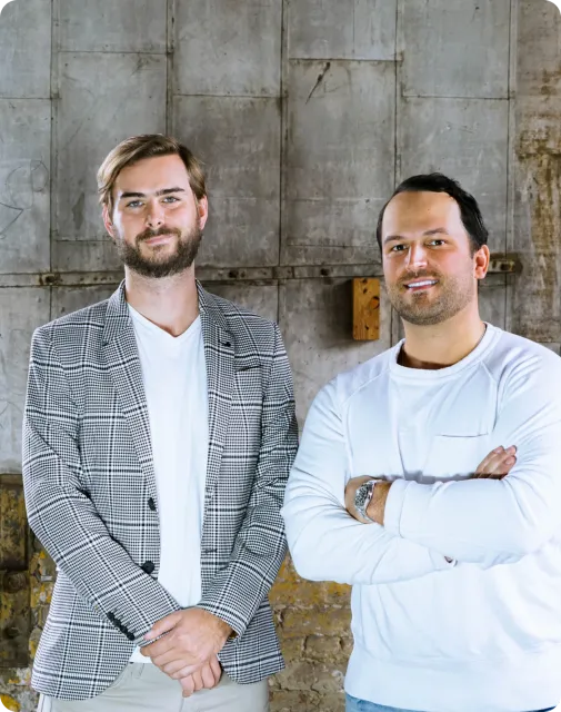 Two men stand side by side in front of a textured, industrial wall, their style as sharp as solar innovation—one in a plaid blazer, the other in a white long-sleeve shirt with arms crossed.
