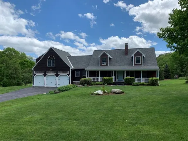 Dark brown house with three-car garage, dormers, and porch, set on a green lawn under a blue sky.