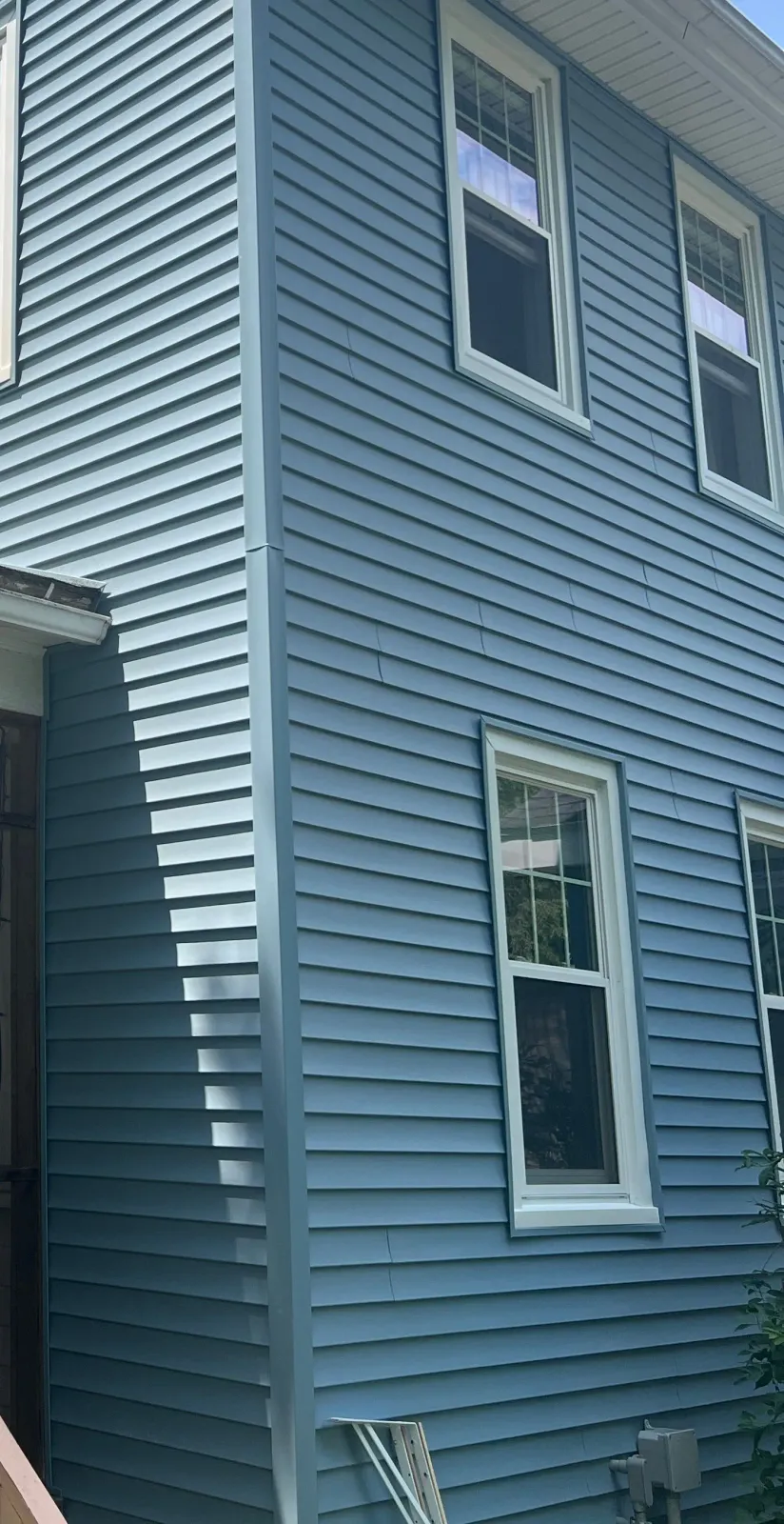 Blue-sided house with white-framed windows. Corner of house angled with vertical trim, sunny day.