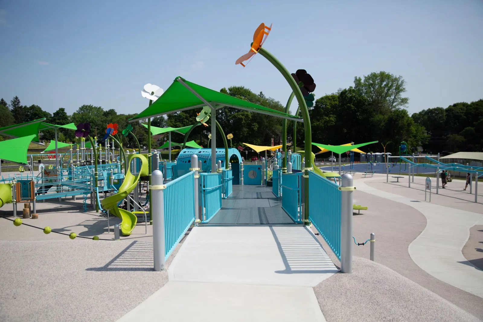 Colorful playground with slides, tunnels, and shade canopies surrounded by a fence and trees in the background.