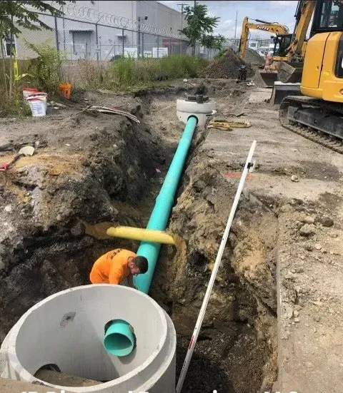 Construction worker installing a large green pipe into an underground trench with construction equipment nearby.