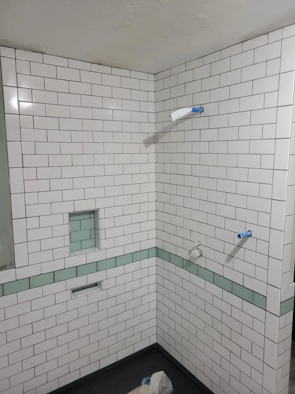 Tiled bathroom shower corner with white subway tiles, green accent band, and two blue pipe fittings in the wall