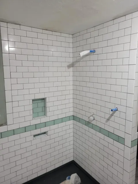 Tiled bathroom shower corner with white subway tiles, green accent band, and two blue pipe fittings in the wall