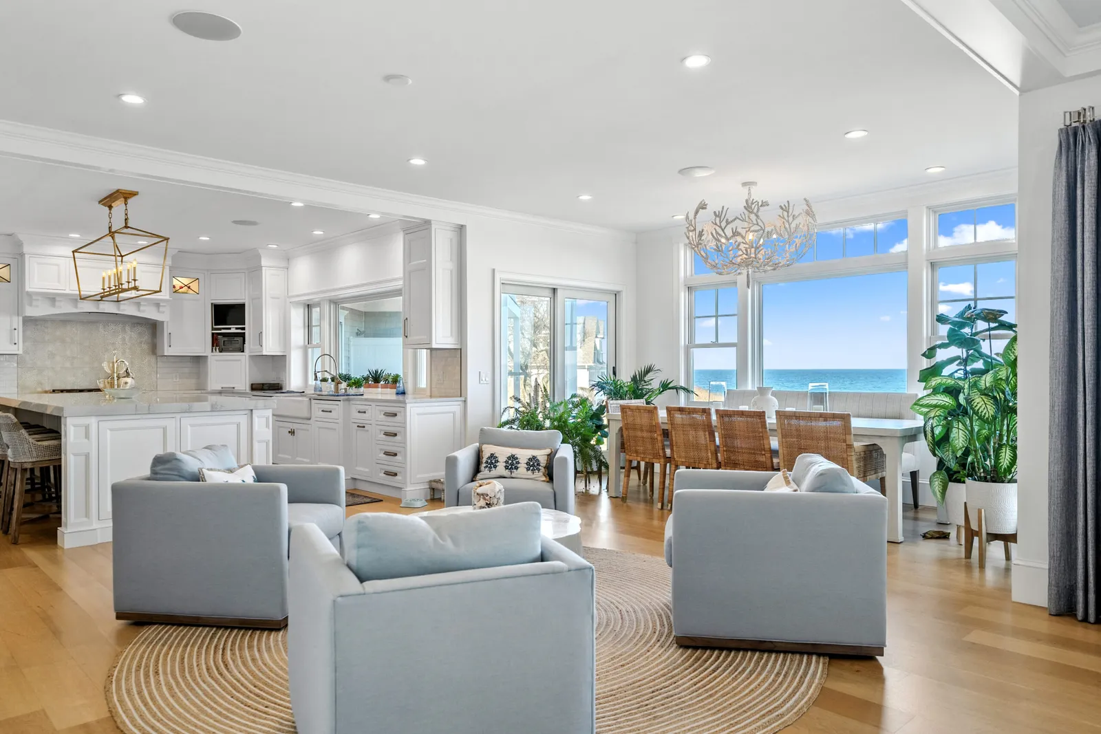 Open-plan living and kitchen area at the Main Street project in Dennis, MA, with light blue sofas, gold chandelier, white cabinetry, wicker dining chairs, and panoramic ocean views