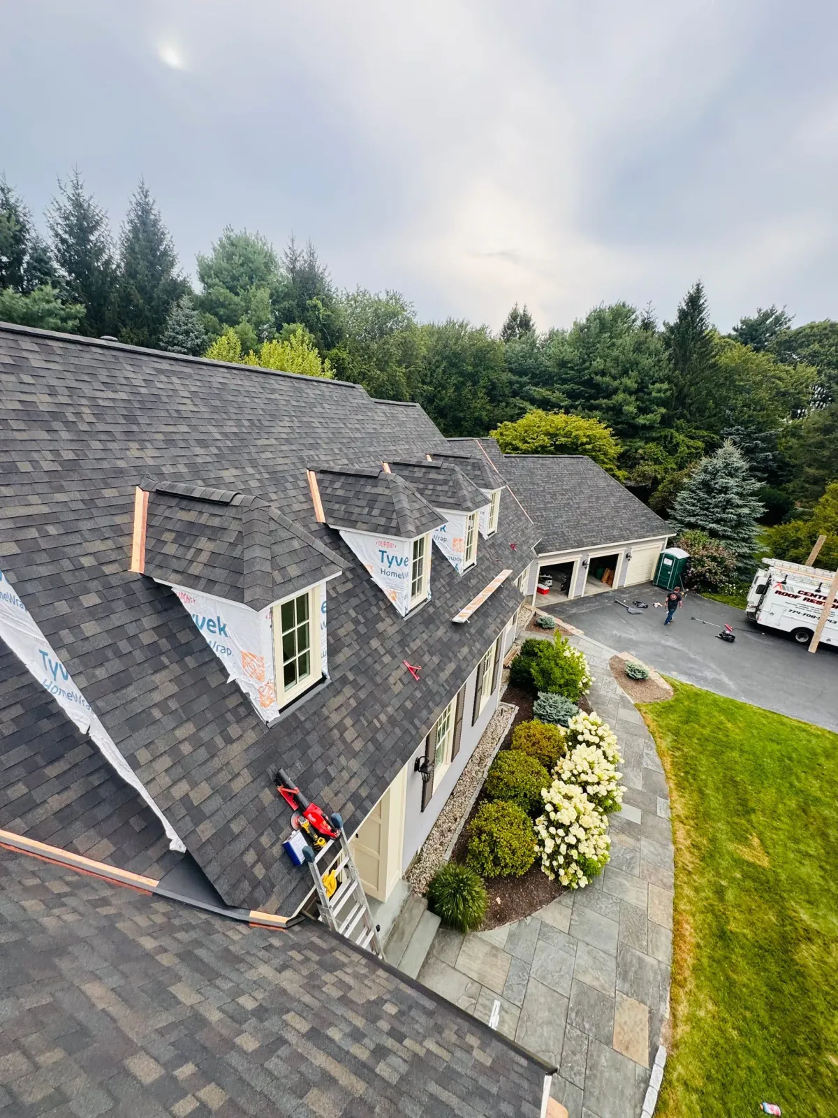 Aerial view of a gray-shingled house with dormers, landscaped yard, and driveway beside a wooded area