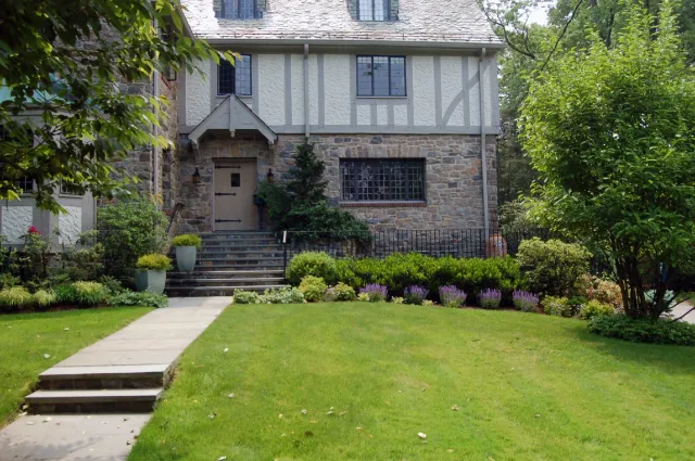 Front view of a house with stone and Tudor-style siding, surrounded by well-maintained lawn and garden with colorful bushes, trees, and potted plants, and stone steps leading to the front door.