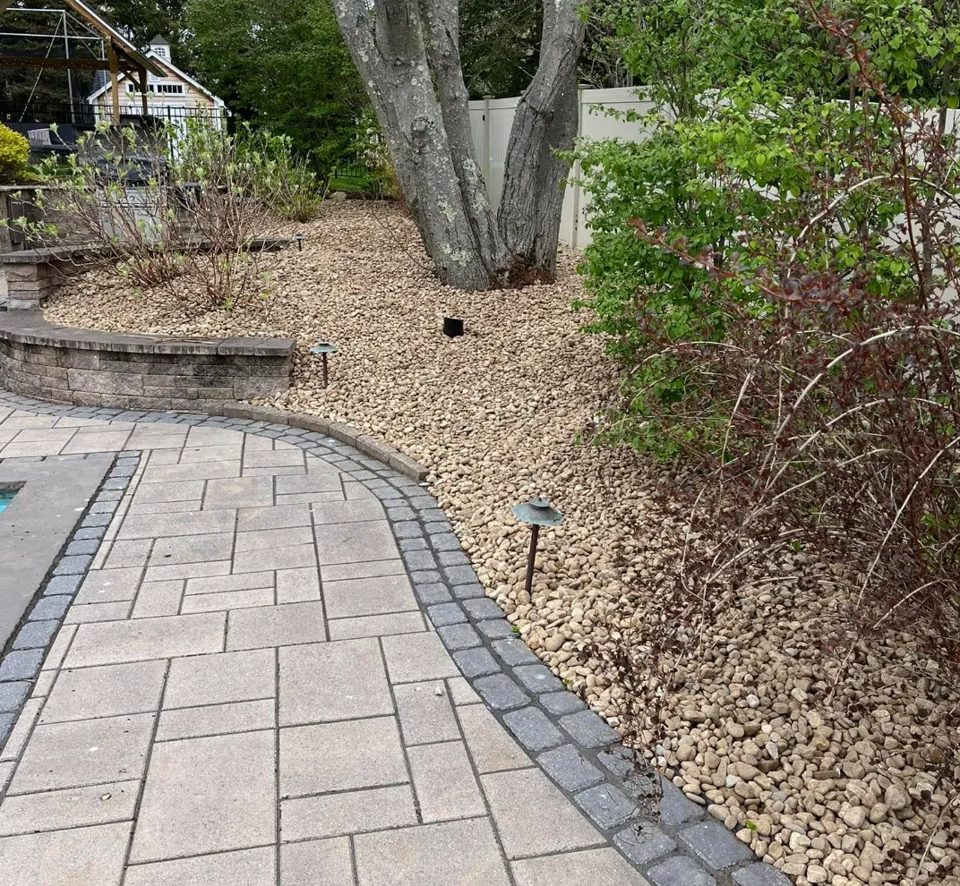 A landscaped backyard with a paved walkway, brick edging, a large tree, bushes, and small ground lights.