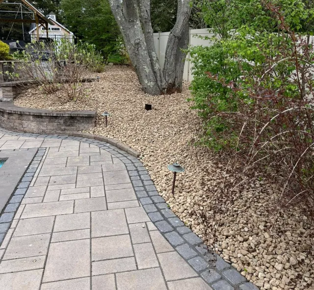 A landscaped backyard with a paved walkway, brick edging, a large tree, bushes, and small ground lights.