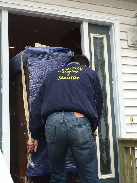 Moving worker in blue jacket and jeans carrying wrapped furniture through a door.