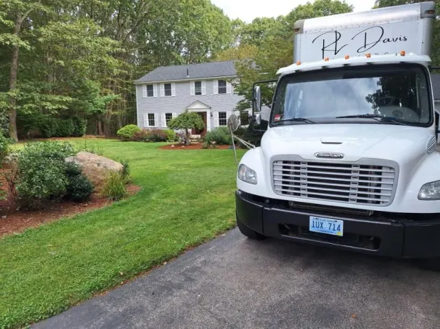 White moving truck parked in front of a gray house with a green lawn.