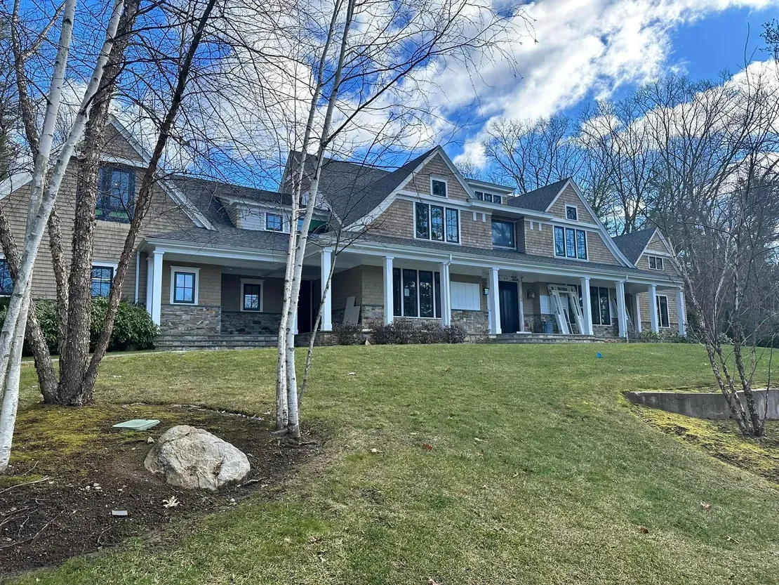 Large house with a covered porch, stone facade, and dormers on a grassy hill under a cloudy sky.