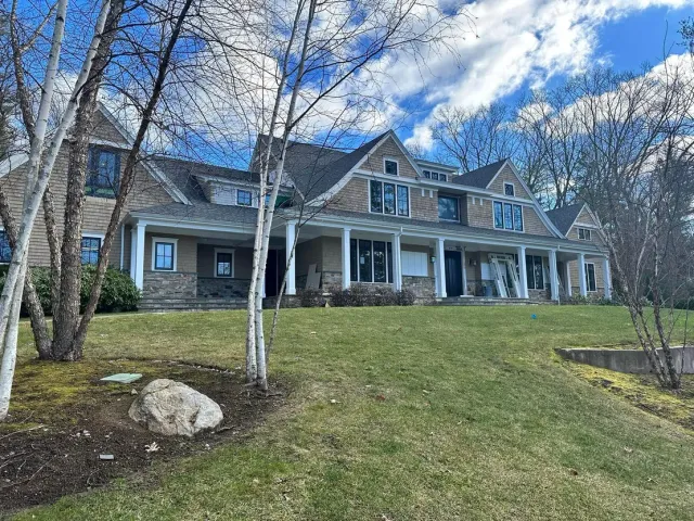 Large house with a covered porch, stone facade, and dormers on a grassy hill under a cloudy sky.