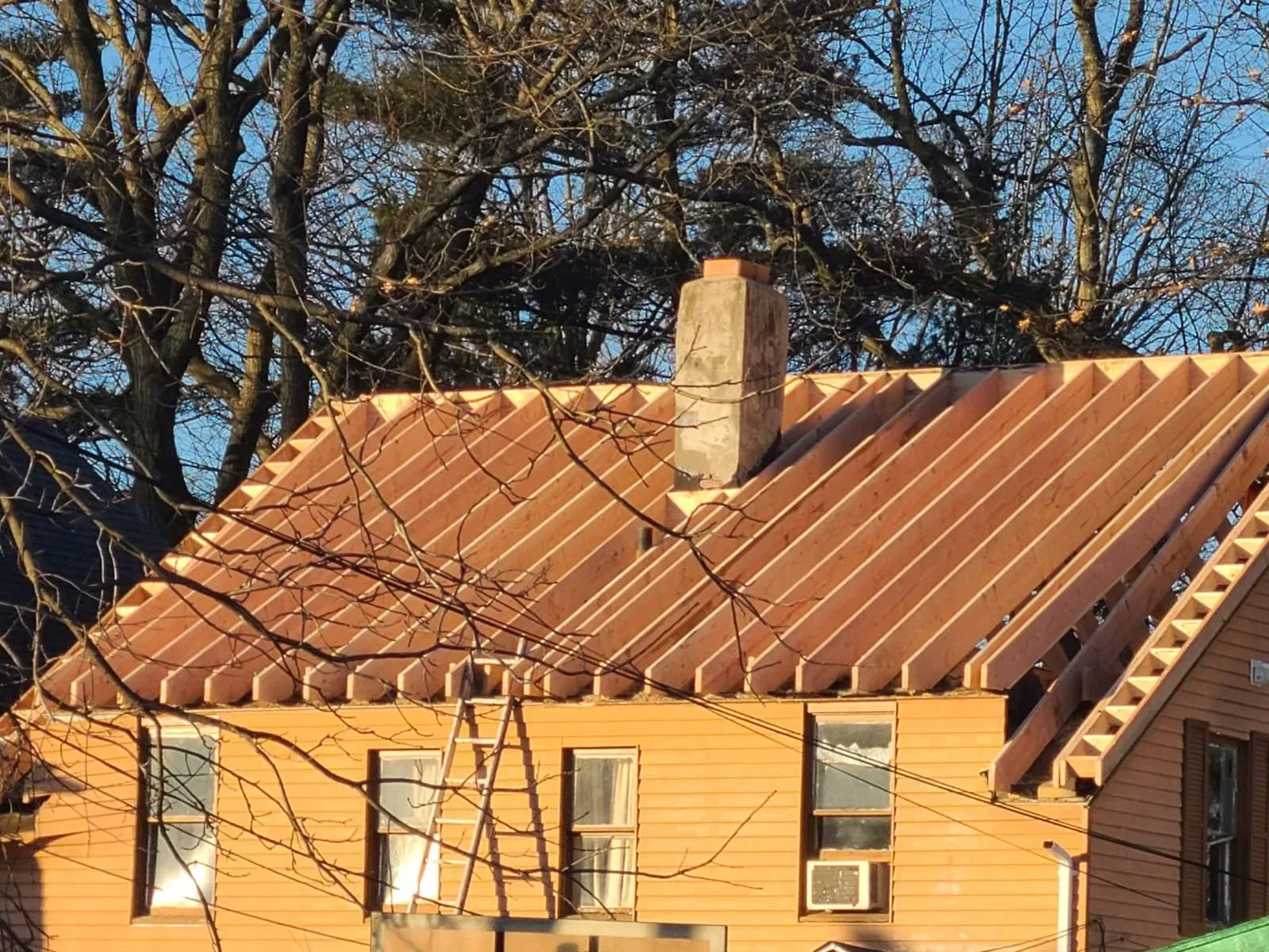 House under construction with exposed wooden roof framing, two visible windows, an air conditioning unit, and a chimney, trees in the background, and a clear blue sky.