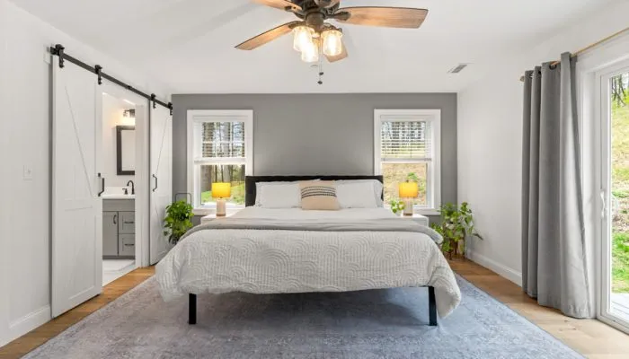 Serene bedroom with king bed dressed in white quilted bedding, gray accent wall behind headboard, white barn-style sliding doors opening to ensuite bathroom, ceiling fan with wood blades, and large windows letting in natural light over hardwood floors.