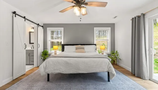 Serene bedroom with king bed dressed in white quilted bedding, gray accent wall behind headboard, white barn-style sliding doors opening to ensuite bathroom, ceiling fan with wood blades, and large windows letting in natural light over hardwood floors.