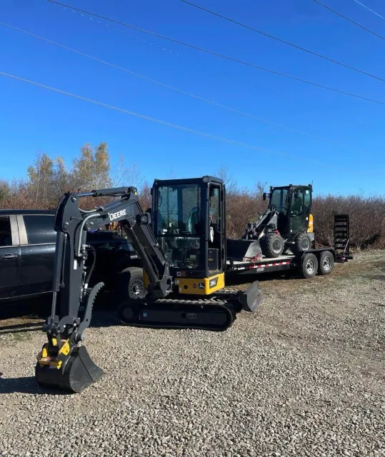A black trailer with two small construction vehicles, a skid-steer loader and an excavator, parked on a gravel lot under a clear blue sky.