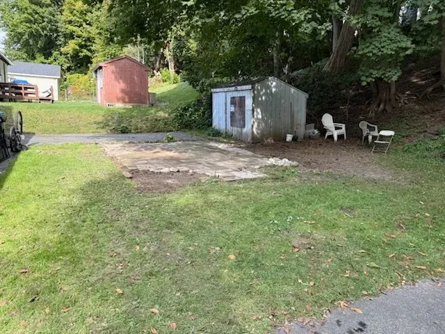 A backyard area featuring a grass lawn, a stone patio, two sheds, and patio chairs near a tree-lined hill.