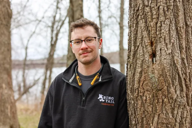 A man with glasses and a beard standing outdoors in front of a large tree, wearing a black jacket with a logo that says 'Ring Tail Carpentry' and yellow ear protection around his neck.