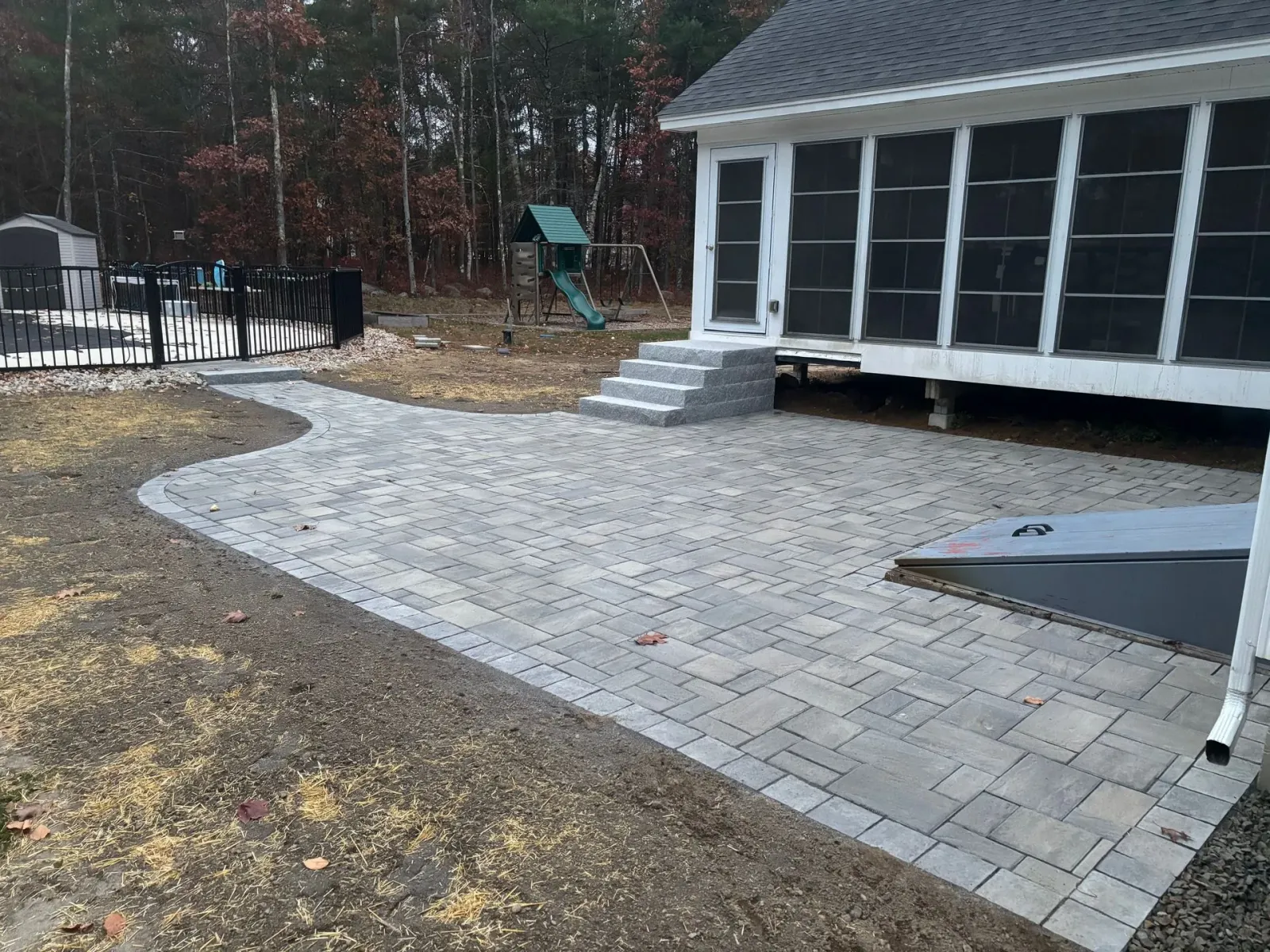 Brick patio next to a house with screened porch and stairs, with a pathway to a fence and playground in the yard.