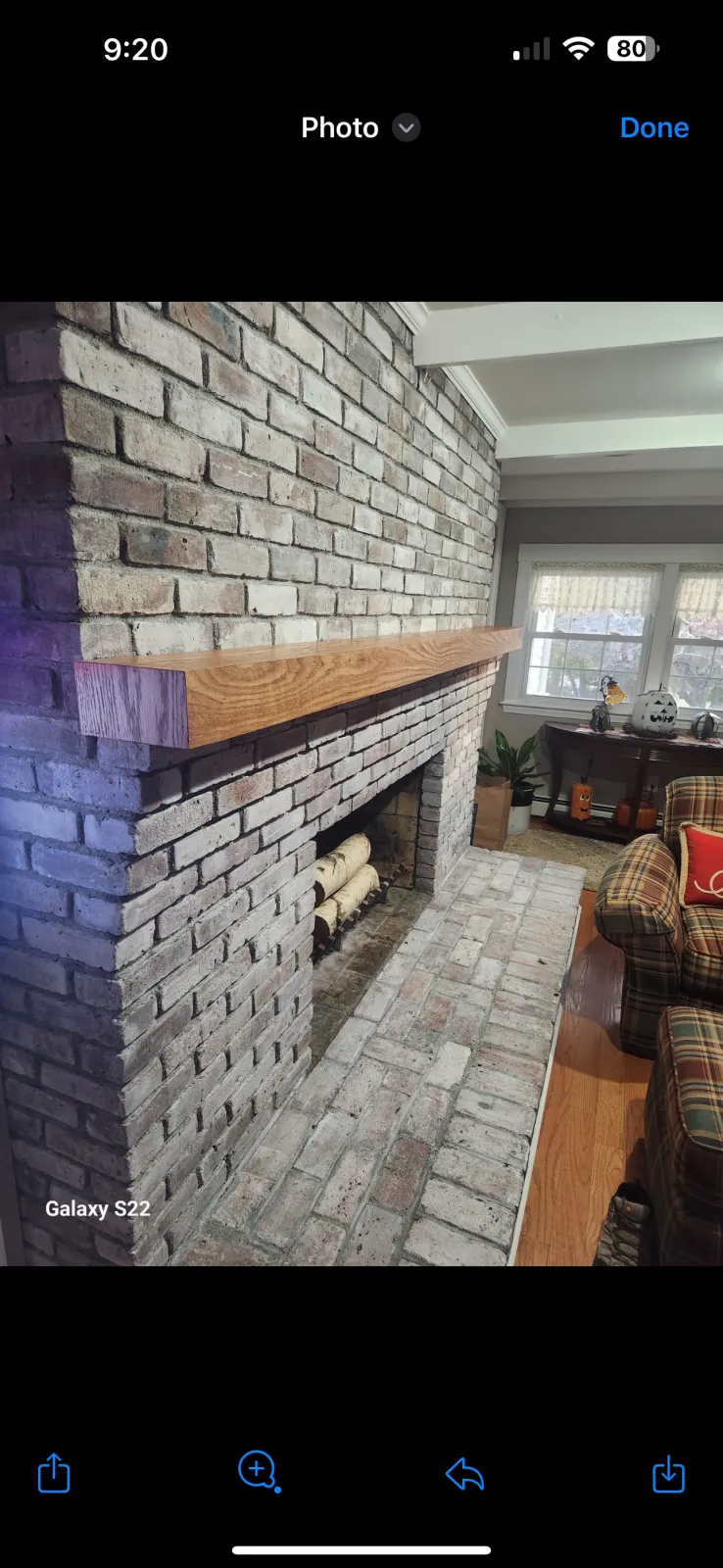 Interior of a house featuring a brick fireplace with a wooden mantel, a window with curtains, a plaid armchair, and a patio table with decorations.