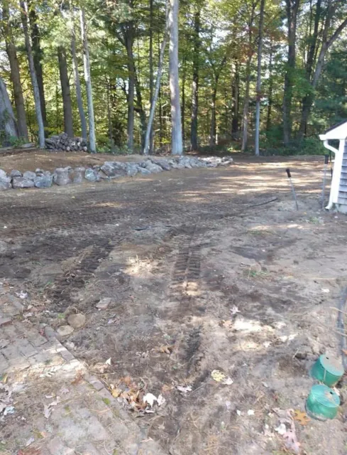 A backyard with dirt ground and tire tracks, surrounded by trees. There is a pile of logs on the left and a row of large rocks on the ground. Part of a house with blue siding is visible on the right.
