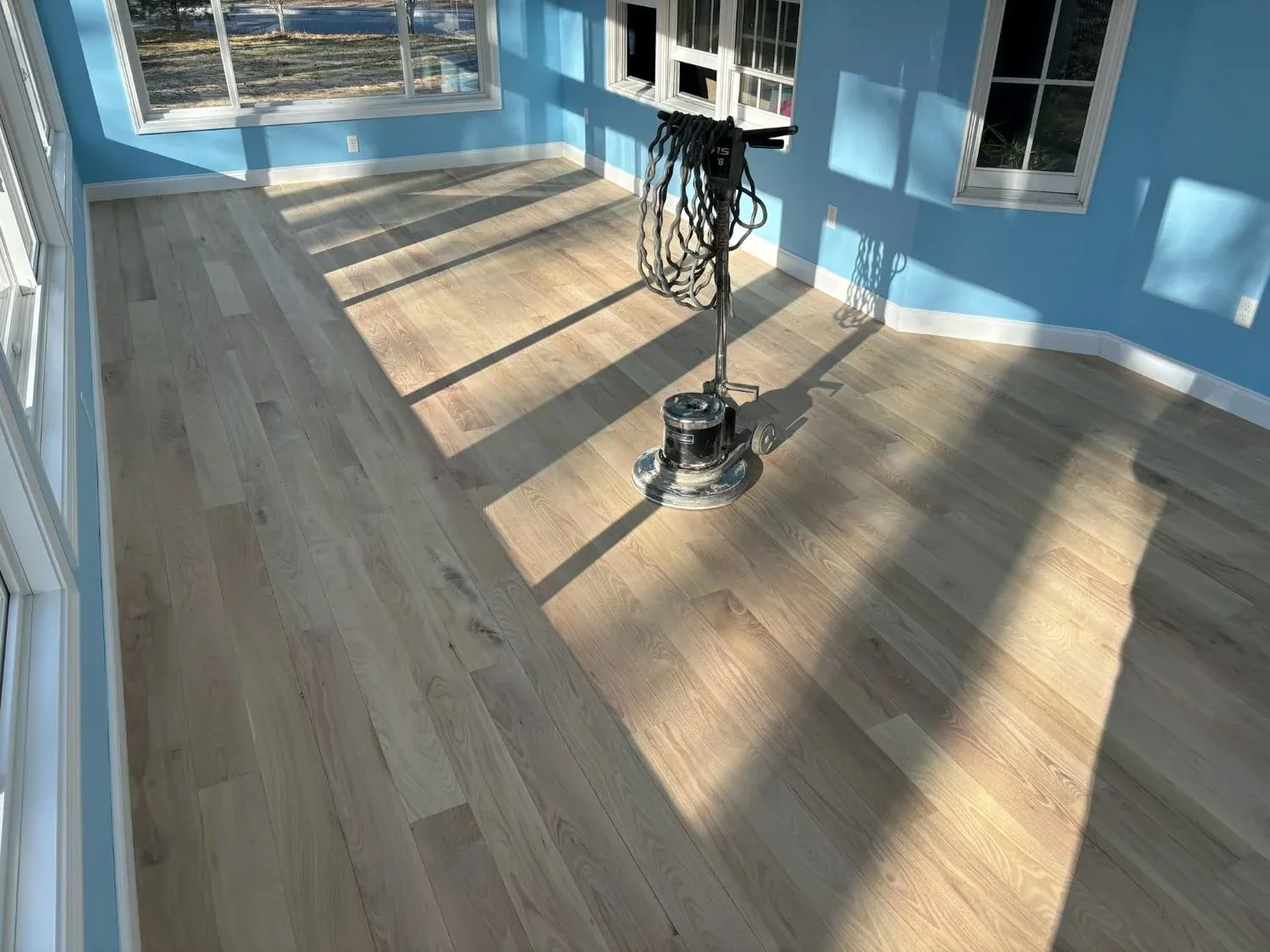 sunroom with light wood floors that have been sanded, blue painted walls, and the electric sander in the middle of the floor
