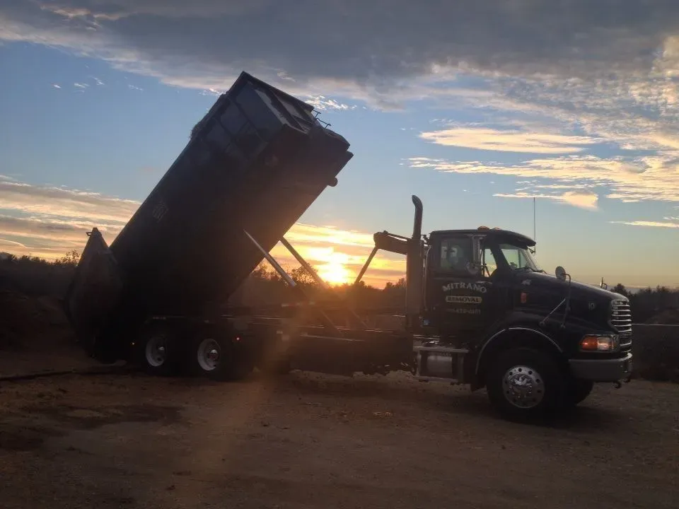 Black dump truck unloading on a dirt lot at sunset.