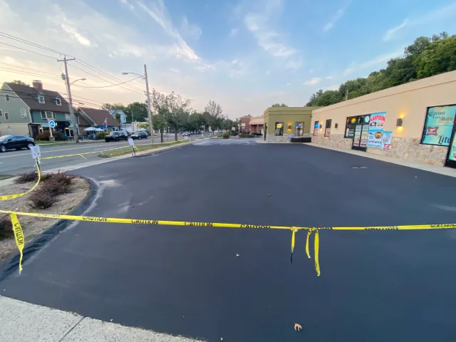 A parking lot section with yellow caution tape across the entrance, which is freshly paved and empty, with a row of storefronts on the right and residential houses and cars on the left under a partly cloudy sky.