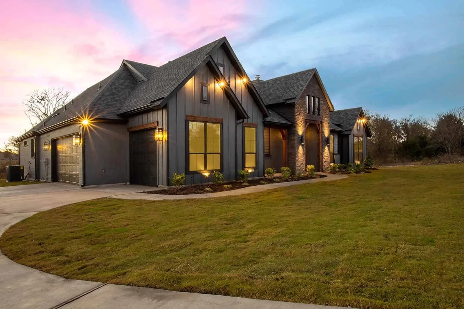 Exterior lighting shown on a dar gray house at dusk with green grass, concrete driveway & mulch beds and flowers.