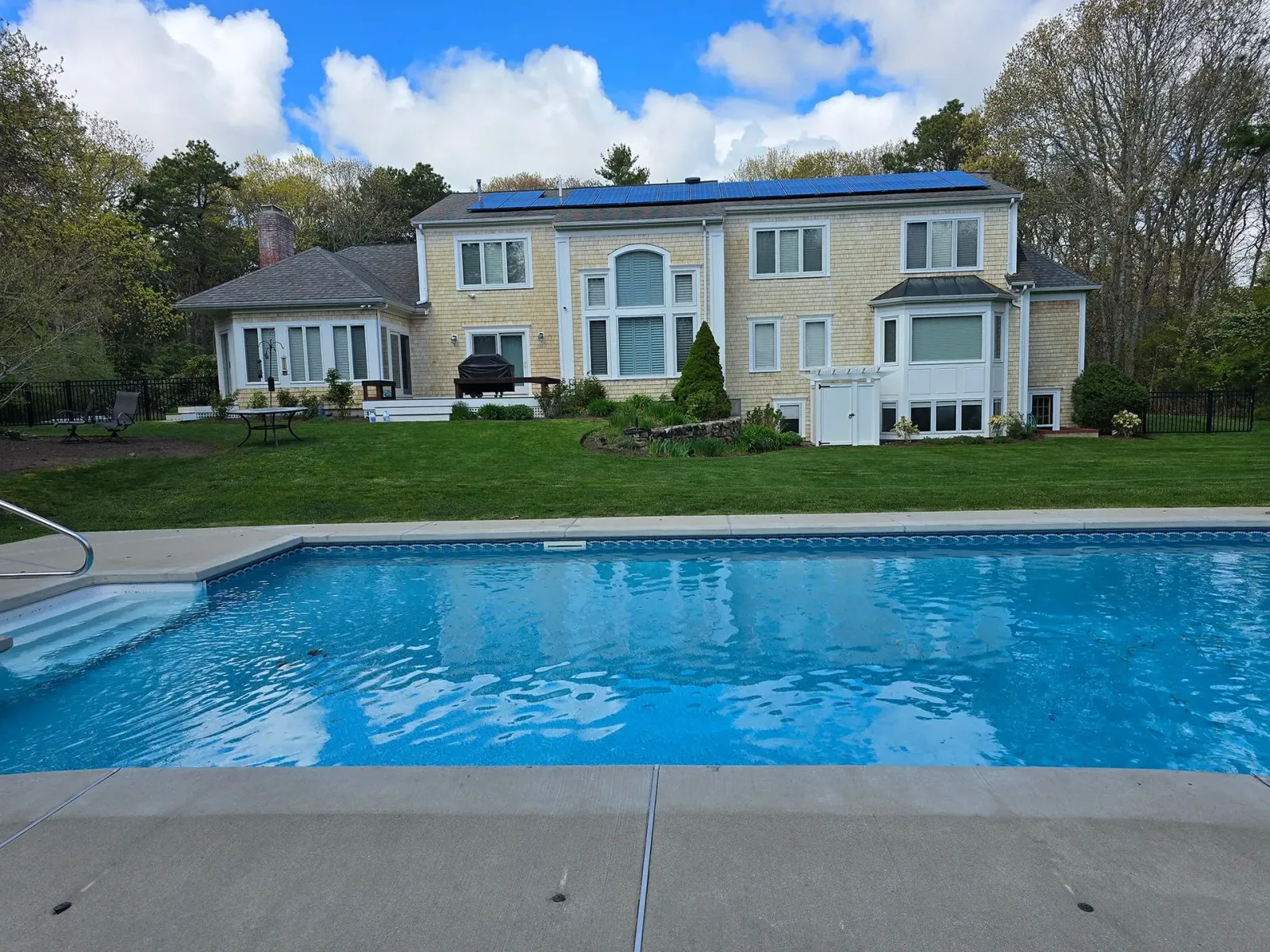 Large two-story beige house with a blue pool in the foreground under a partly cloudy sky