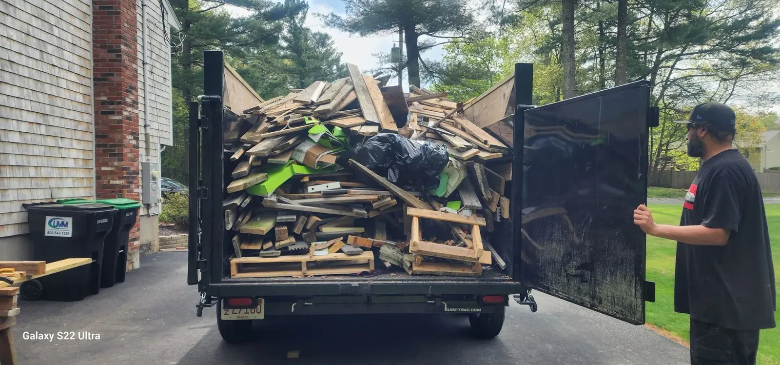 A man closing the tailgate of a truck filled with wood debris, parked on a driveway near a brick building.