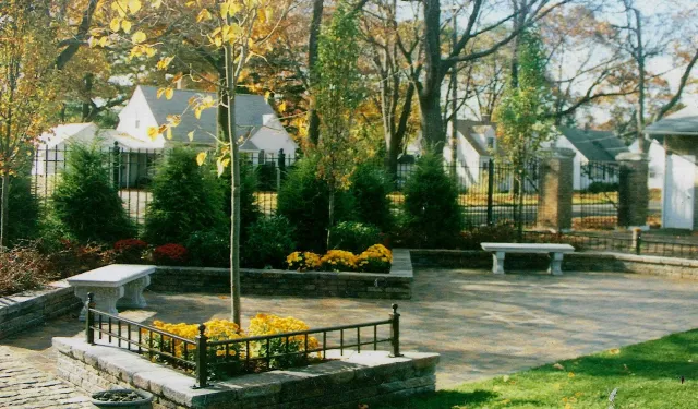 Park with stone benches, trees, and flower beds, bordered by a wrought iron fence and brick walls; houses in the background.