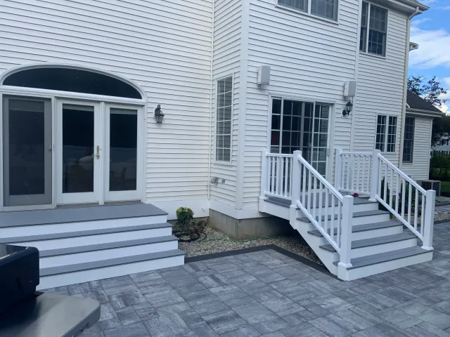 Backyard with white stairs leading to sliding glass doors and a deck, stone patio, and beige siding house