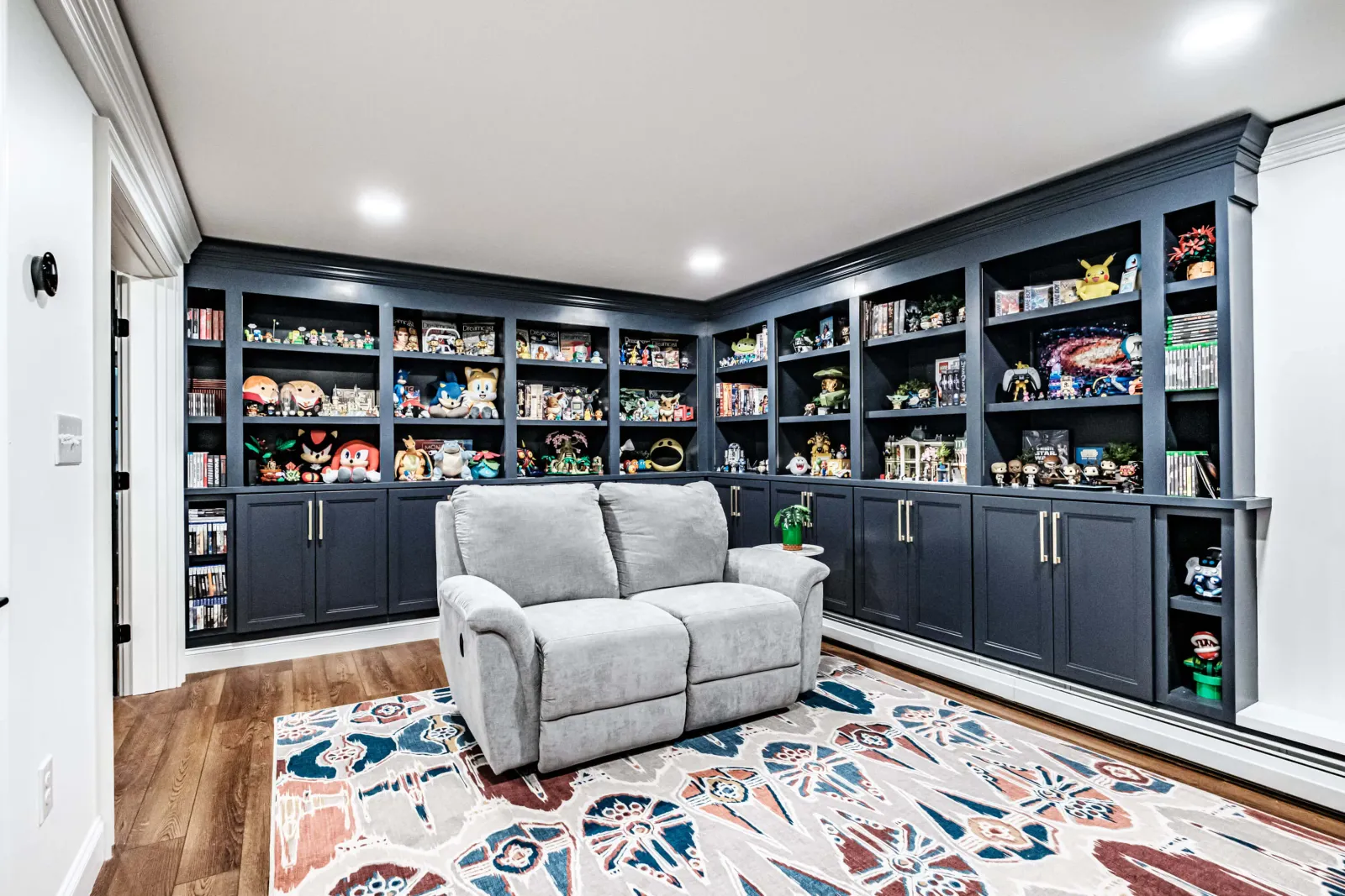 Gray reclining loveseat on a patterned rug in front of dark built-in shelves filled with collectibles.
