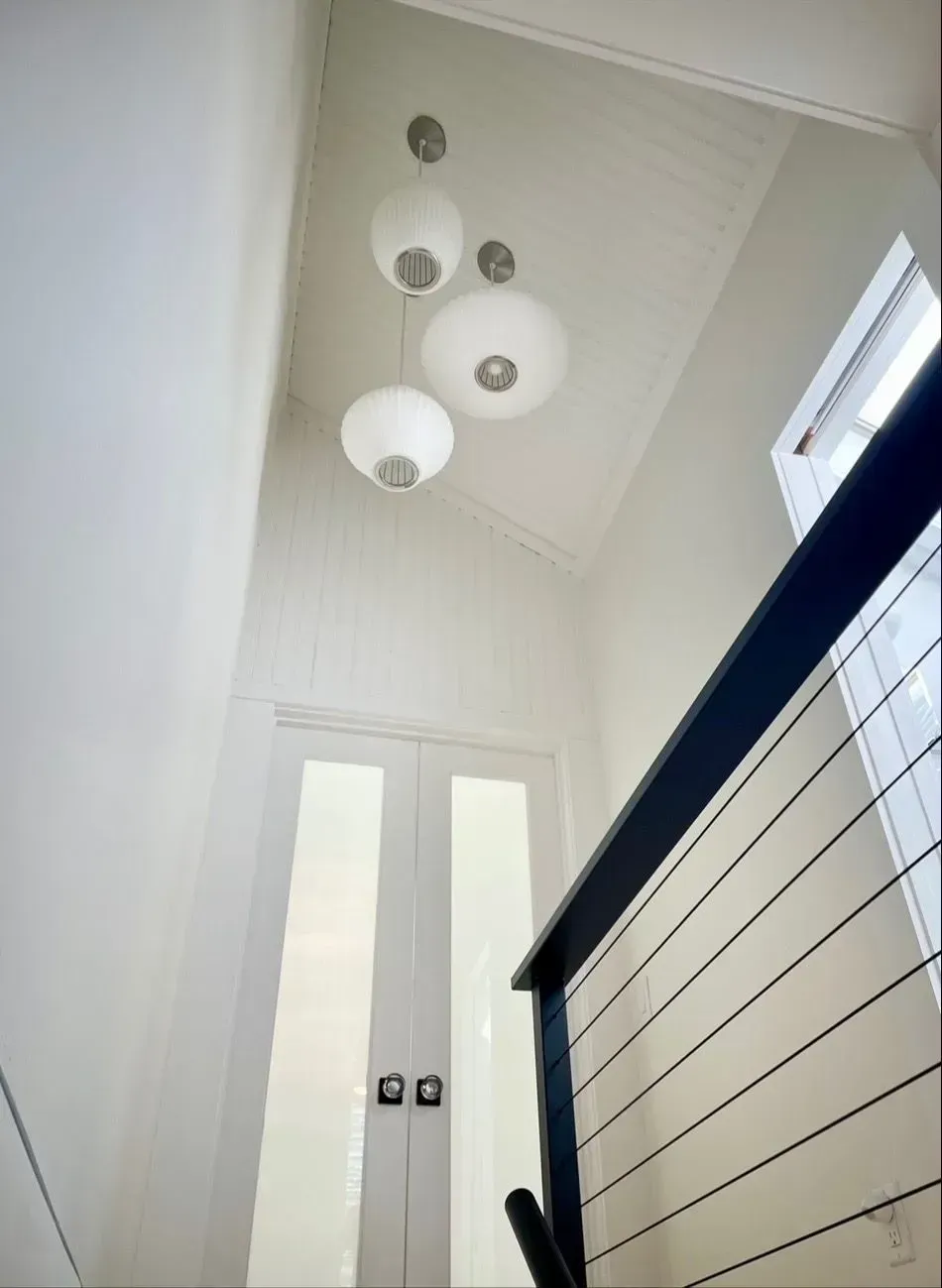 Bright stairwell with white walls, pendant lights, and a black handrail beside tall windows.