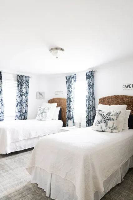 A bedroom with white bedding, starfish-themed pillows, wicker headboards, blue and white patterned curtains, and a ceiling light fixture.