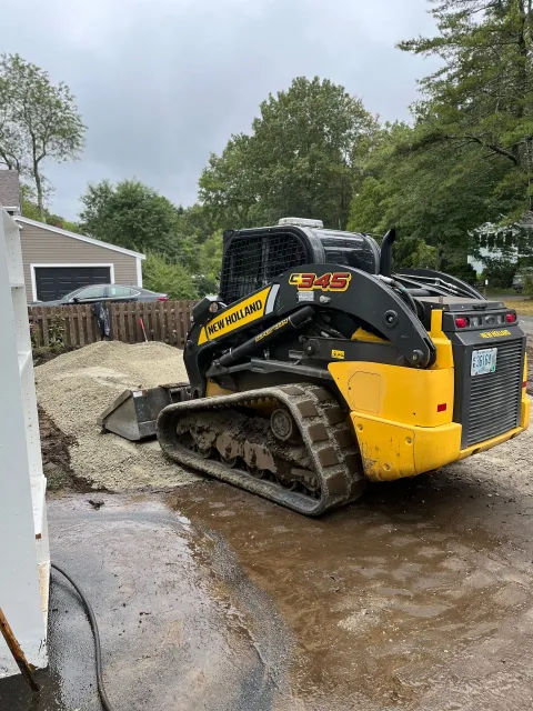 Yellow skid steer loader next to gravel pile, parked on a wet driveway.